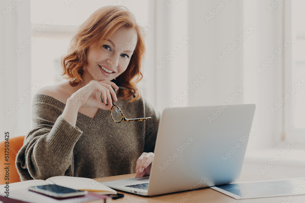 Pleased female scientist makes research on laptop computer, poses in coworking space, searches information for course work, wears warm sweater, prepares for academy test with modern technologies Pleased female scientist makes research on laptop computer, poses in coworking space, searches information for course work, wears warm sweater, prepares for academy test with modern technologies