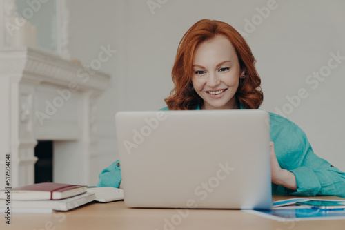 Focused cheerful redhead successful businesswoman does research, makes business project, sits in front of laptop computer at home office desk, uses software application, has happy smile on face
