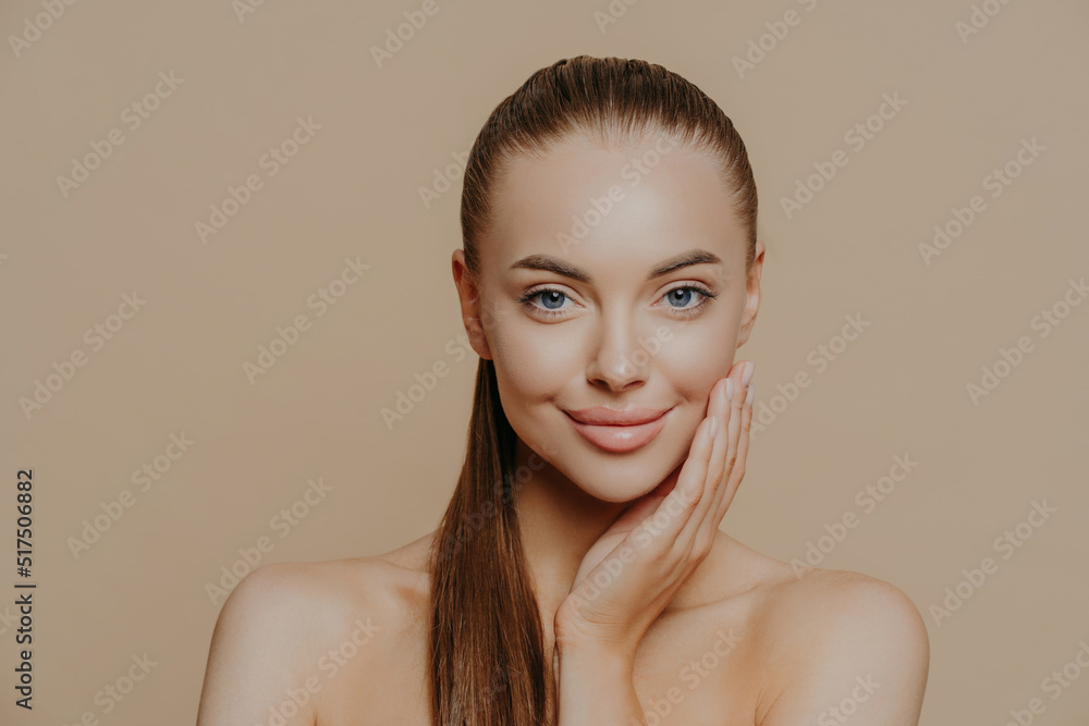 Photo of lovely young woman has smooth healthy skin after beauty procedures and receiving facial massage, stands bare shoulders, has long dark straight hair, isolated over beige wall cares of her body Photo of lovely young woman has smooth healthy skin after beauty procedures and receiving facial massage, stands bare shoulders, has long dark straight hair, isolated over beige wall cares of her body