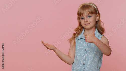 Toddler toddler girl showing thumbs up pointing empty place, advertising area for commercial text, copy space for goods promotion. Young little child kid. Studio shot indoors on pink wall background