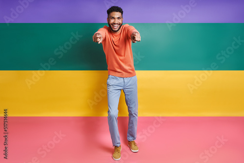 Full length of happy African man pointing camera while standing against colorful background