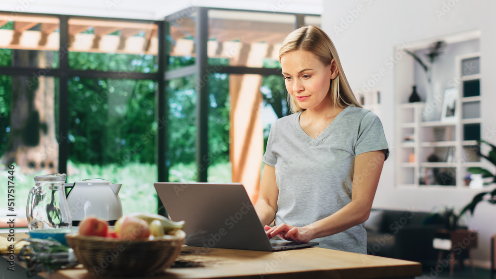 Portrait of Beautiful Young Adult Woman with Blond Hair Wearing Gray V-Neck T-Shirt, Using Laptop Computer while Standing in Living Room. Successful Woman Working from Home in Bright Apartment. Portrait of Beautiful Young Adult Woman with Blond Hair Wearing Gray V-Neck T-Shirt, Using Laptop Computer while Standing in Living Room. Successful Woman Working from Home in Bright Apartment.