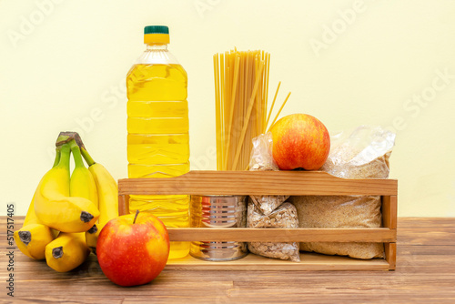 Fototapeta Naklejka Na Ścianę i Meble -  Donation food box, volunteering help,humanitarian aid full of canned,sunflower oil, pasta,cereal,grain and apples,banana,fruits. Delivery wooden box for charity on yellow, wooden background still life