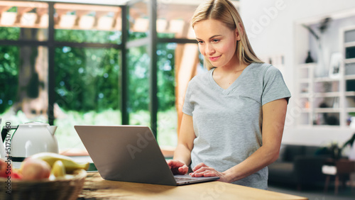 Portrait of Beautiful Young Adult Woman with Blond Hair Wearing Gray V-Neck T-Shirt, Using Laptop Computer while Standing in Living Room. Successful Woman Working from Home in Bright Apartment.