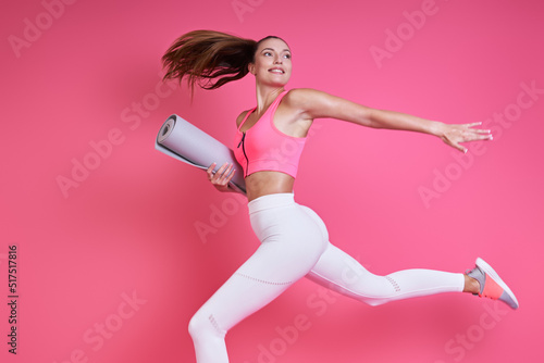 Fototapeta Naklejka Na Ścianę i Meble -  Beautiful woman in sports clothing carrying an exercise mat while running against pink background