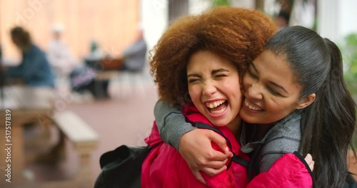 Cheerful best friends embracing and having fun while standing in a mall or at college. Two young multiethnic LQBTQ women laughing, hugging, and sharing a kiss on the cheek while meeting to catch up