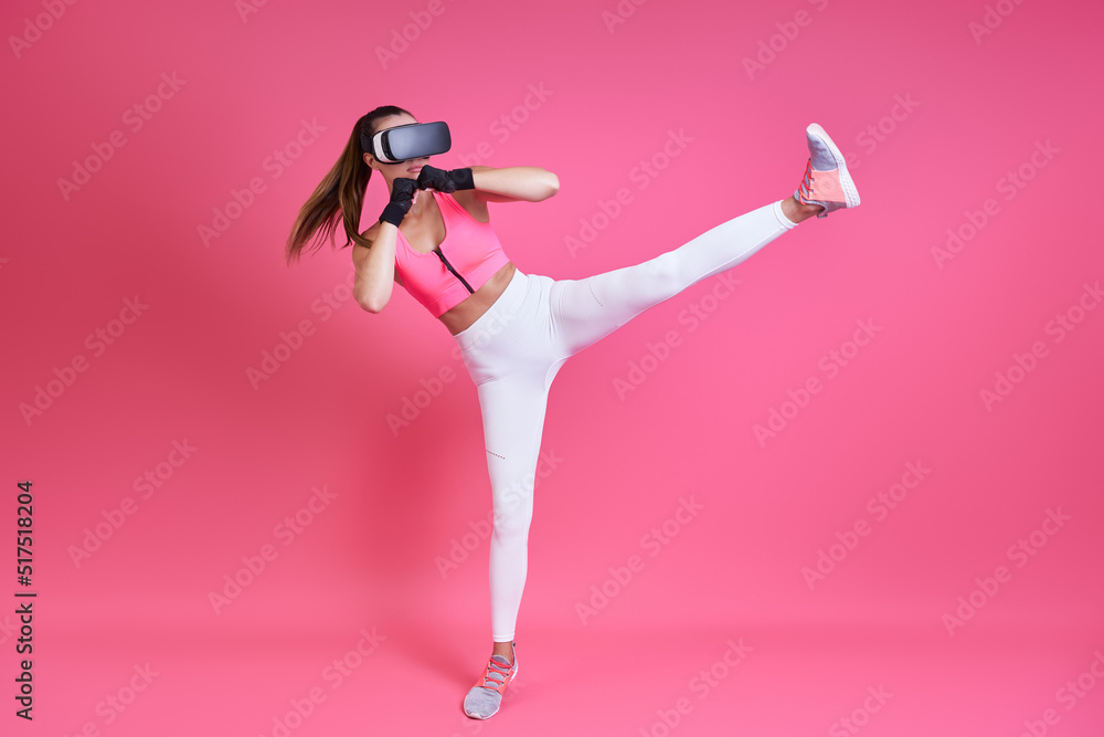 Young woman in virtual reality glasses kicking against pink background