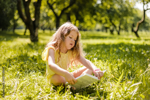 A little girl is sitting with a book in the garden. A girl reads a book on a summer day © Sviatlana