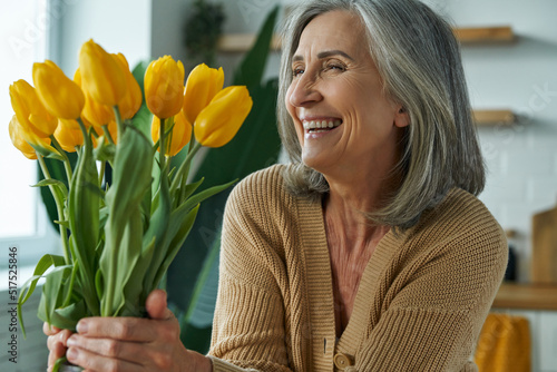 Elegant senior woman holding a bunch of yellow tulips and smiling while relaxing at home #517525846