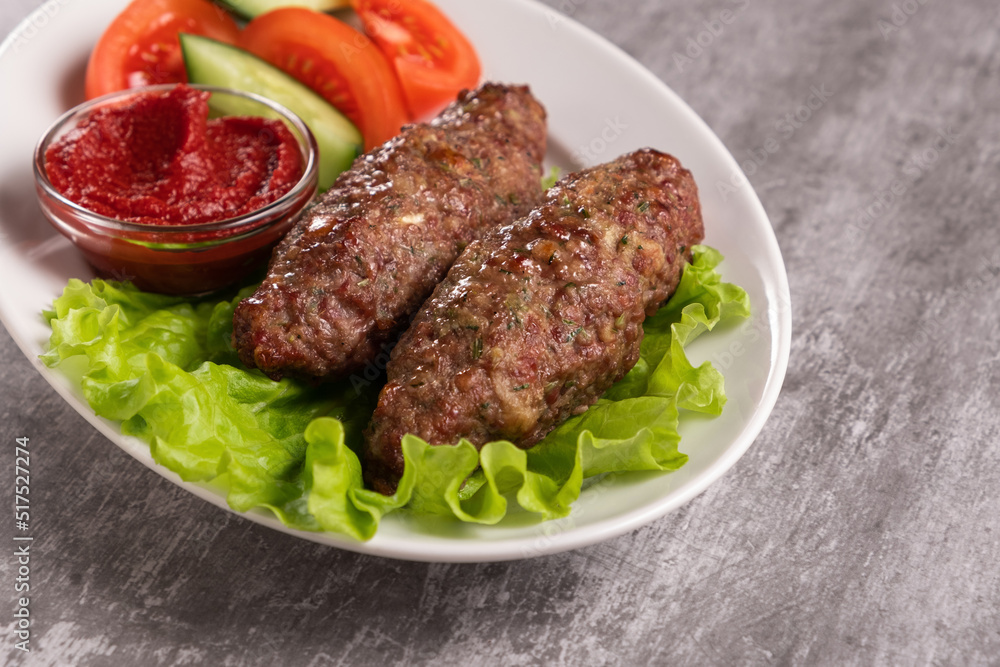 The national dish lyulya-kebab with vegetables and lettuce leaves with red sauce in a plate on a gray background. Horizontal orientation, close-up, no face, copy space