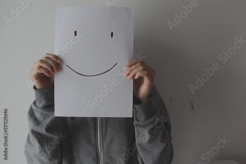 Woman holding a smiling face emoticon in front of face against a gray background, depression, mental health, masking feelings.