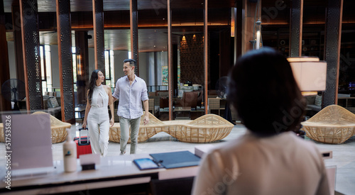 Young couple walking in lobby and arriving at hotel reception while front desk employee welcoming and greeting them