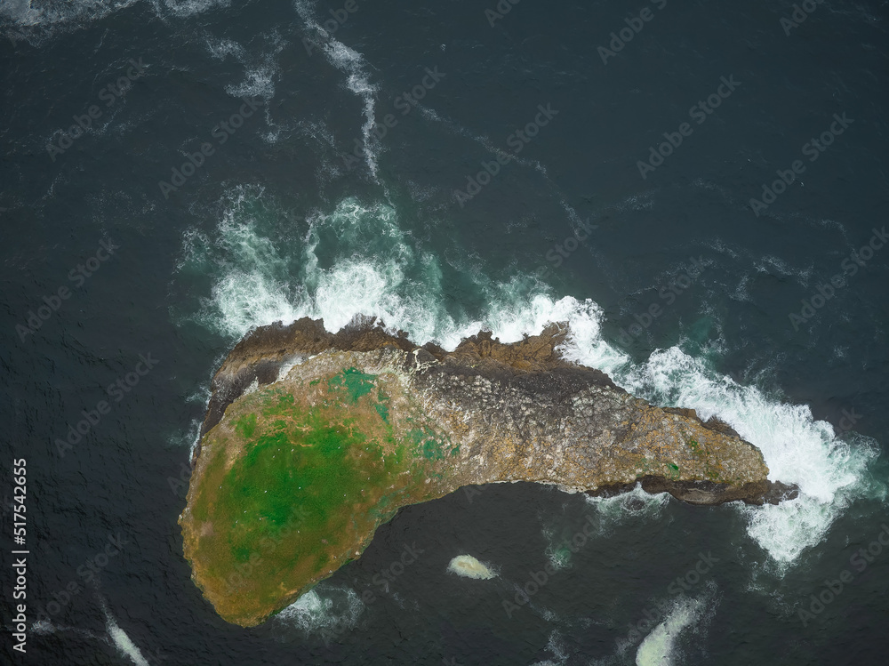 Aerial view. A stone island in the ocean that looks like a big fish. In places overgrown with green moss. Dark turquoise water and white foamy waves. Advertising of tourist destinations.