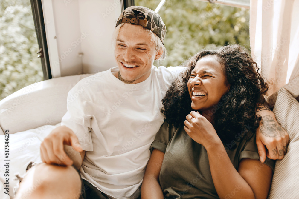 Fototapeta premium Mixed-race couple chilling on white sofa, laughing and hugging, enjoying their relationship's vibes, African woman feeling happy sitting next to her Caucasian hipster man, both dressed casually
