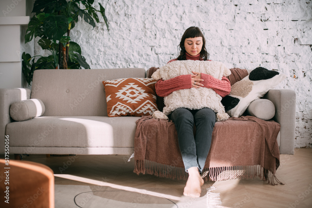 Obraz premium A young woman sits comfortably on a sofa in a modern bright apartment. A beautiful brunette girl in a knitted sweater is relaxing at home