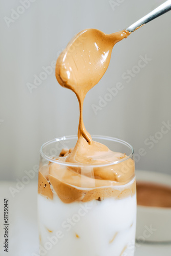 Vaso de Café Dalgona, Delicioso café coreano, café frío, cuchara sirviendo más crema al vaso, con fondo blanco
