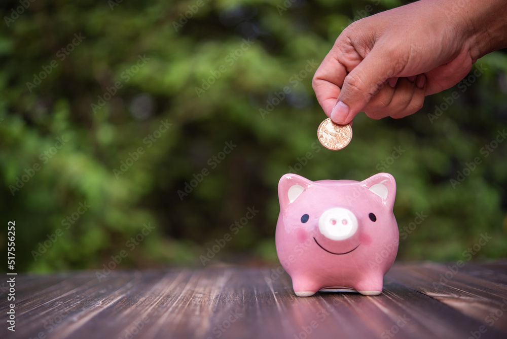 hand put a coin into piggy bank on wooden table with green natural leaves background