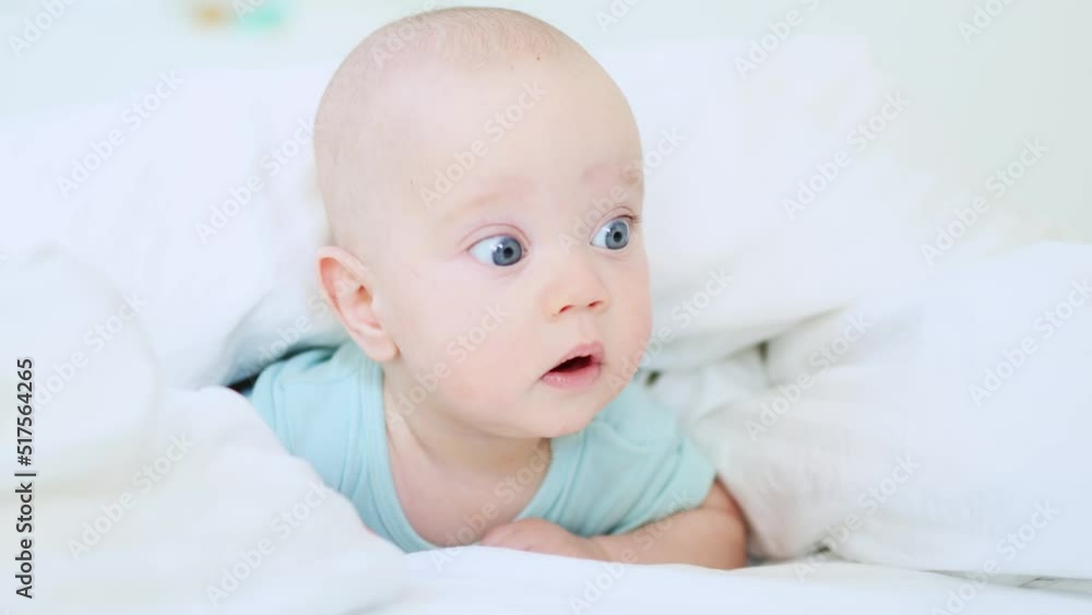funny caucasian baby infant lying in white bed on stomach in the morning, happy 3 month child in blue clothes awake in sunny day bedroom, smiling portrait of blue-eyed kid in white bedsheets funny caucasian baby infant lying in white bed on stomach in the morning, happy 3 month child in blue clothes awake in sunny day bedroom, smiling portrait of blue-eyed kid in white bedsheets