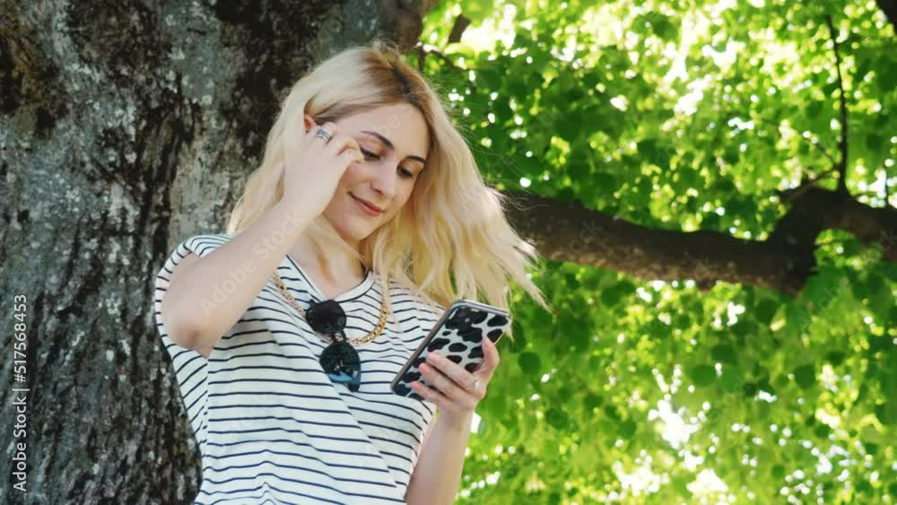 young cheerful girl sitting close to a tree and reading something pleasant in her phone. medium shot outdoor. High quality 4k footage