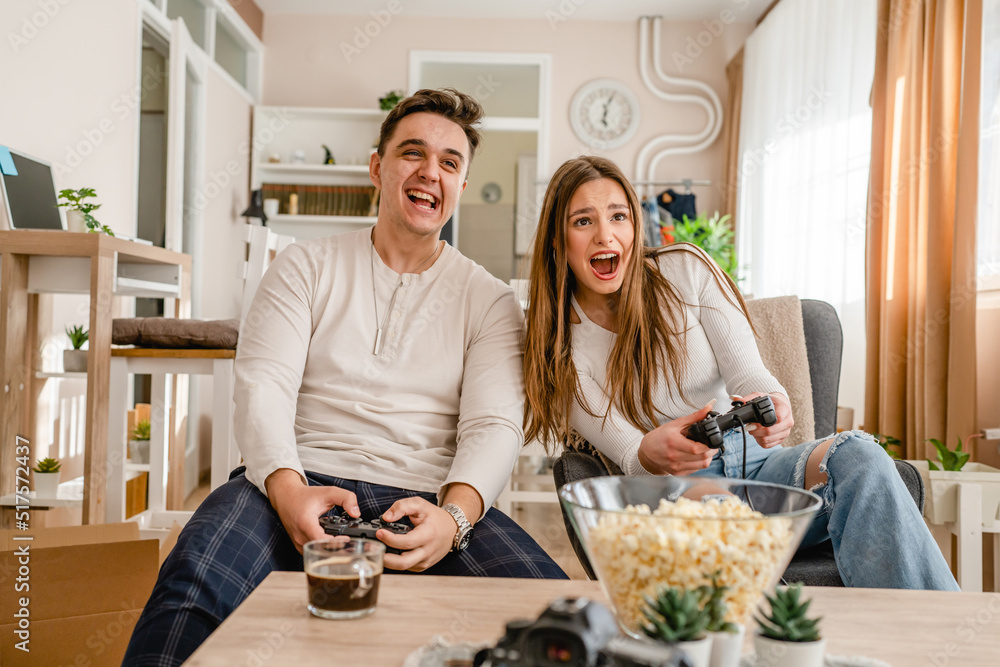 two caucasian people sitting at home playing video game console having fun young couple man and woman boyfriend and girlfriend spending time together holding joystick controller smiling front view