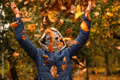 Defocus Halloween people. Person in grim reaper mask raising hand and throwing leaves. Many flying orange, yellow, green dry leaves. Out of focus