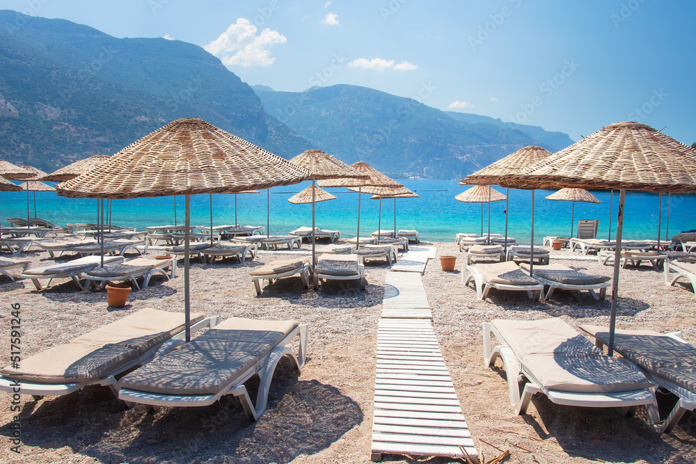 Landscape of beautiful coastline in Oludeniz with straw umbrellas. View from beach on blue sea and mountains, amazing rest in Turkey. Beginning of day on beach without people.