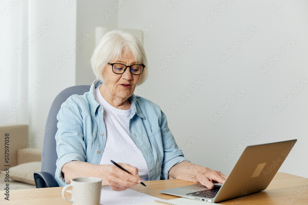 portrait of an elderly woman in a light shirt working at a desk through a laptop at home, carefully looking at the monitor. The concept of working from home