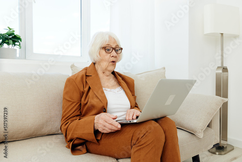 an emotional, surprised elderly woman is sitting at home in a brown suit holding a laptop on her lap and staring at the monitor with her mouth wide open trying to understand the problem