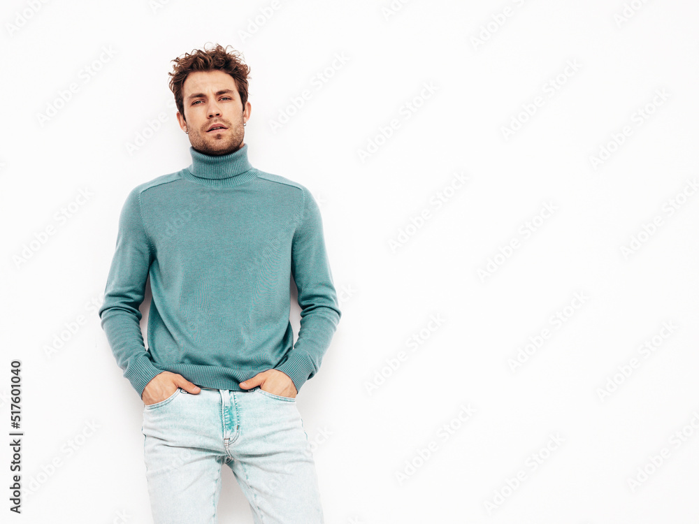 Fototapeta premium Portrait of handsome confident model. Sexy stylish man dressed in blue sweater and jeans. Fashion hipster male with curly hairstyle posing near white wall in studio. Isolated