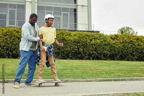 African happy dad pushing his son while he standing on skateboard, he teaching him to ride in park
