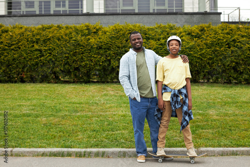 Portrait of African happy boy standing on skateboard and smiling at camera together with his dad while they riding in park