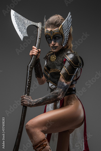 Portrait of wild valkyrie dressed in armor holding huge axe and staring at camera against grey background.