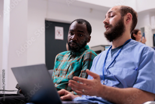 Nurse consulting african american patient in waiting area, using laptop to explain disease diagnosis and recovery treatment in reception lobby. People talking about healthcare at facility.