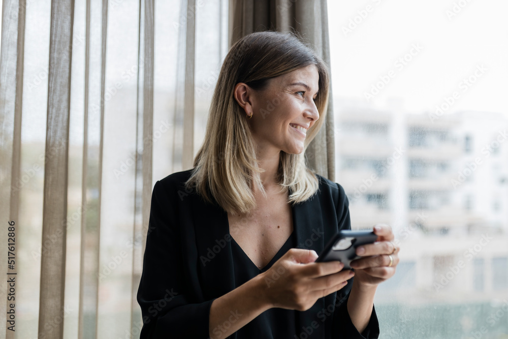Smiling businesswoman holding mobile phone looking through window Smiling businesswoman holding mobile phone looking through window