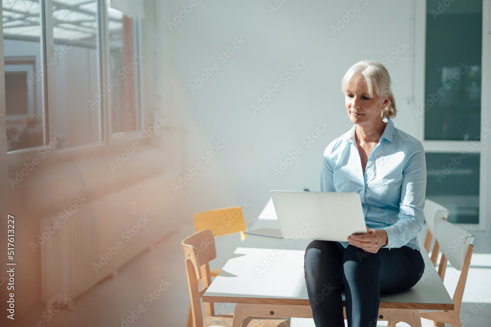 Senior businesswoman using laptop sitting on desk in office Senior businesswoman using laptop sitting on desk in office