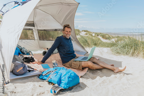 Happy freelancer with laptop sitting in tent at beach on sunny day photo