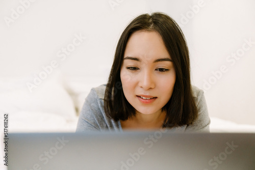 Young concentrated asian girl lying with laptop on bed