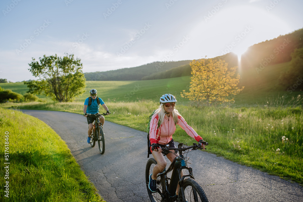 Fototapeta premium Active senior couple riding electric bicycles on road at summer park, healthy lifestyle concept.