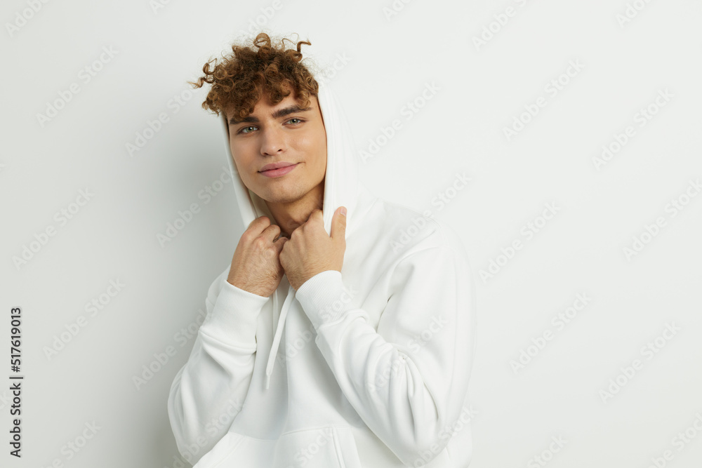 a cute man stands on a white background in a white hoodie and looks pleasantly into the camera adjusting the hood with his hands a cute man stands on a white background in a white hoodie and looks pleasantly into the camera adjusting the hood with his hands