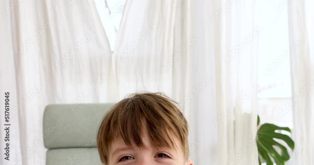 Cheerful boy 5 years old smiles wide hiding in room against bright window. Portrait of schoolboy with long bangs looking in camera closeup Cheerful boy 5 years old smiles wide hiding in room against bright window. Portrait of schoolboy with long bangs looking in camera closeup