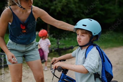 Portrait of young family with little children preapring for bike ride, standing with bicycles in nature, putting on helmets.