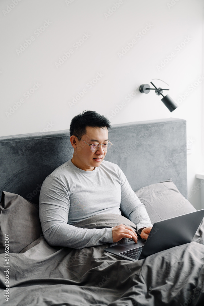 Fototapeta premium Asian man working with laptop after sleep while sitting in bed