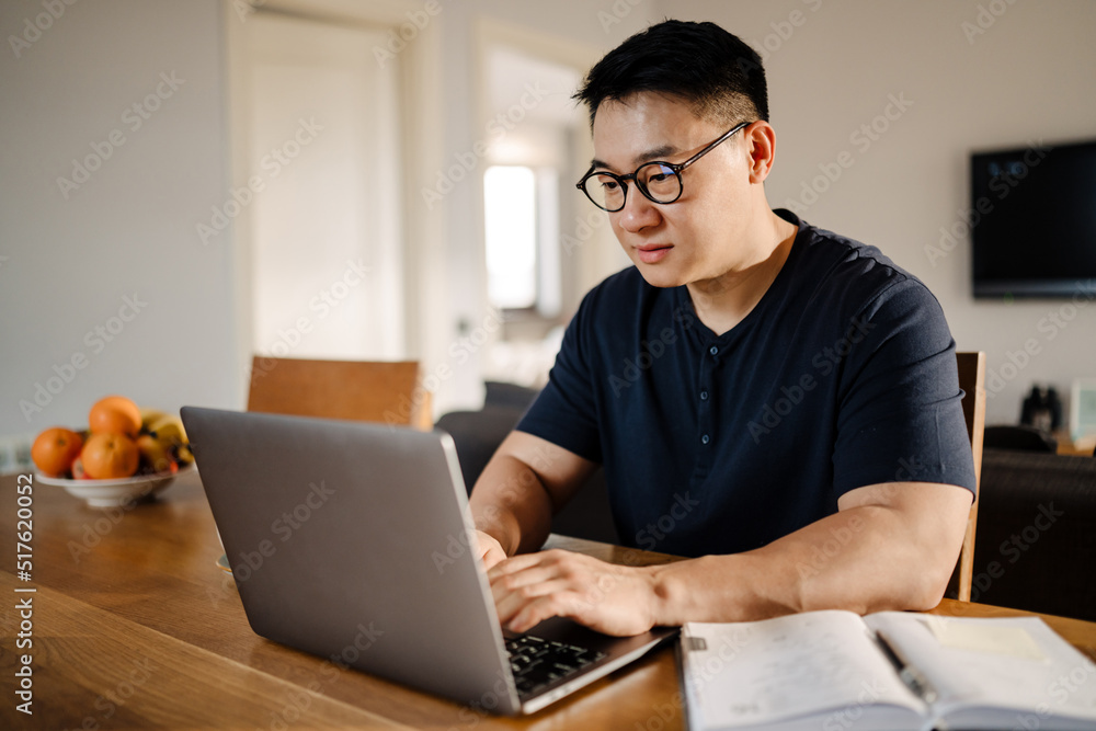 Brunette adult asian man smiling and using laptop computer at home Brunette adult asian man smiling and using laptop computer at home