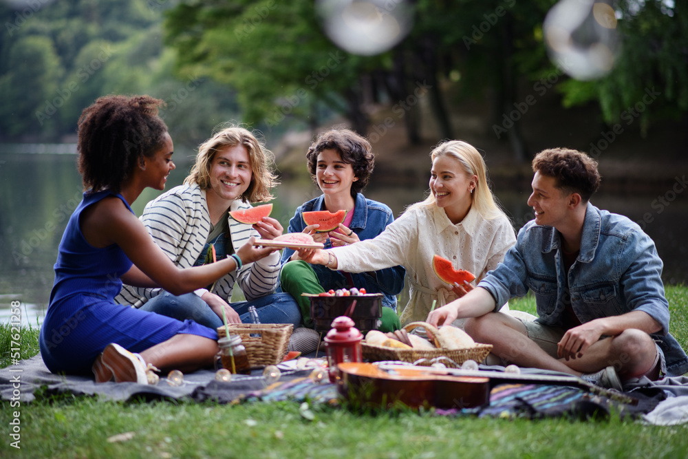 Group of young friends having fun on picnic near a lake, sitting on blanket and eating watermelon.