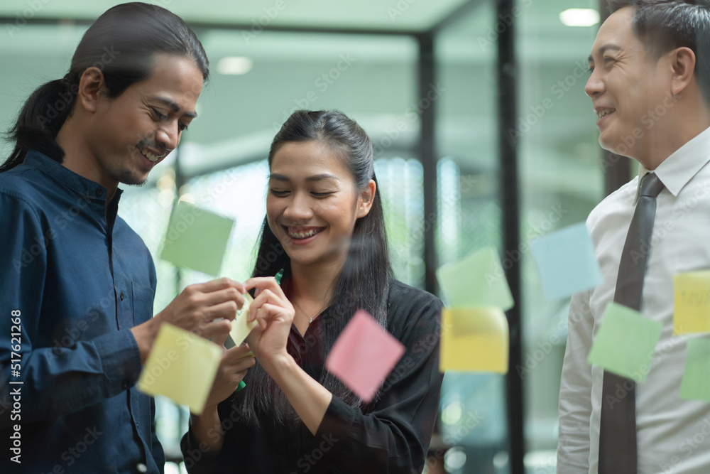 Obraz premium Asian business team meeting in the office using post it notes stick on glass wall. Corporate people brainstorming idea for project together.