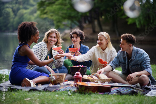 Group of young friends having fun on picnic near a lake  sitting on blanket and eating watermelon.