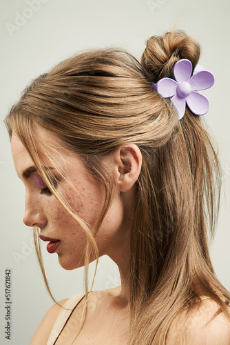 Cropped close-up shot of a blonde girl with a claw clip in her hair. Portrait of a young woman with a violet flower hair claw clip on a pastel background. Side view.