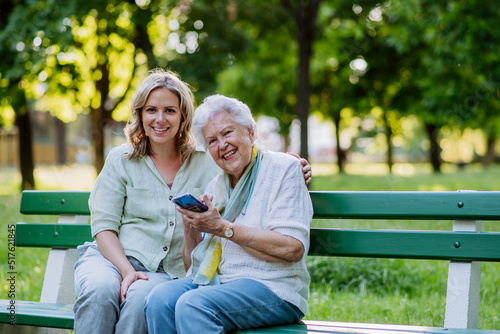 Adult granddaguhter helping her grandmother to use cellphone when sitting on bench in park in summer.