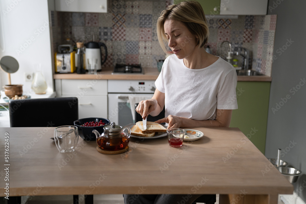 Pensive middle-aged single woman in home wear sit alone at table in kitchen, enjoying breakfast in peace and quiet at home, mature lone lady eating healthy food in morning, spreading butter on toast
