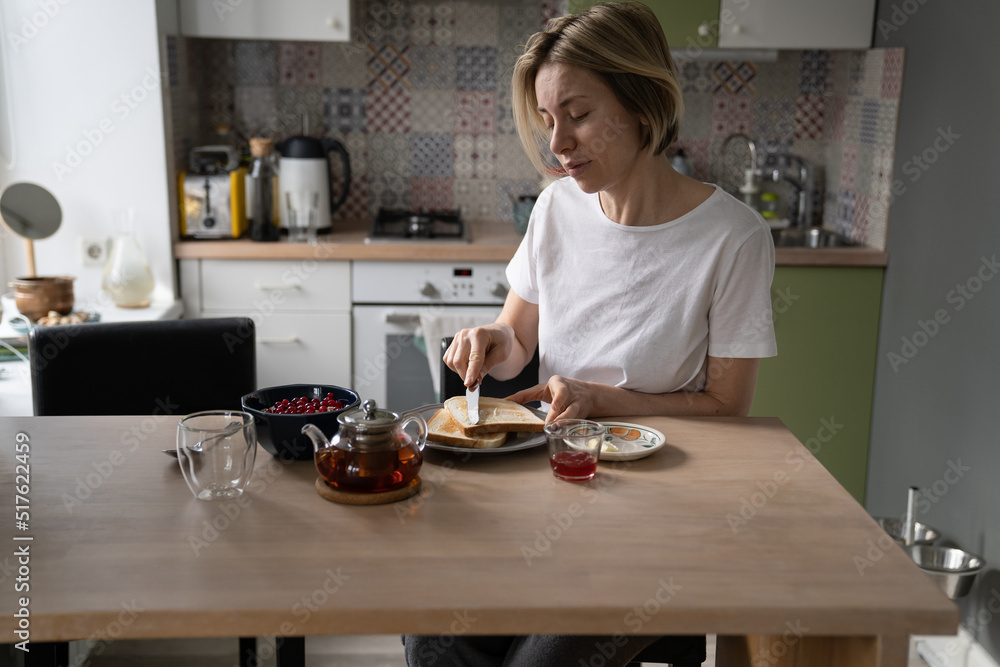 Pensive middle-aged single woman in home wear sit alone at table in kitchen, enjoying breakfast in peace and quiet at home, mature lone lady eating healthy food in morning, spreading butter on toast Pensive middle-aged single woman in home wear sit alone at table in kitchen, enjoying breakfast in peace and quiet at home, mature lone lady eating healthy food in morning, spreading butter on toast